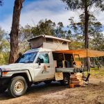 A white 4WD vehicle, equipped with a pop-top roof tent and an orange side-mounted awning, is set up for camping in a sunny forested area, providing shade and an extended outdoor living space.