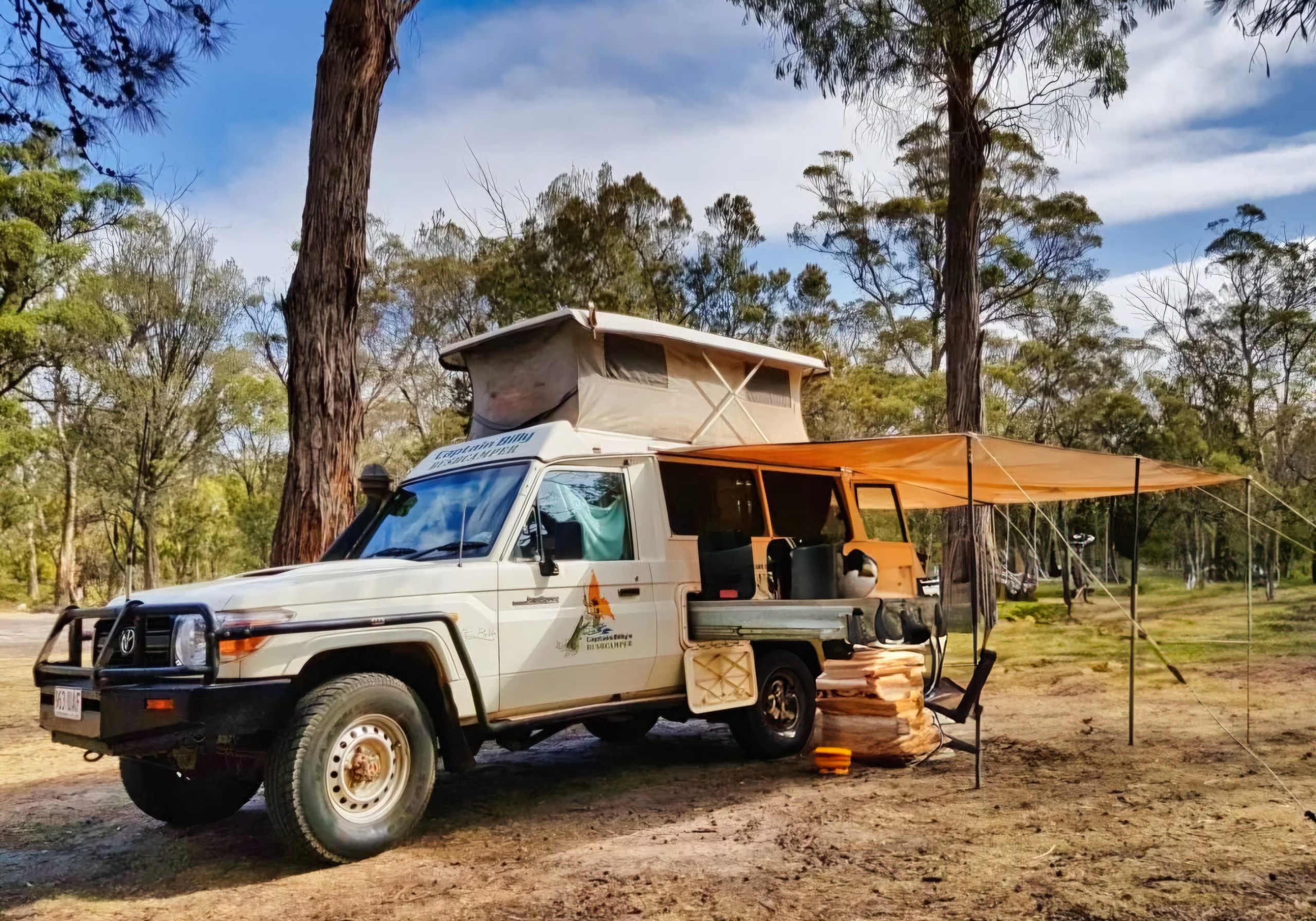 A white 4WD vehicle, equipped with a pop-top roof tent and an orange side-mounted awning, is set up for camping in a sunny forested area, providing shade and an extended outdoor living space.