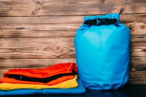 A bright blue dry bag stands upright next to a stack of folded orange, black, and yellow quick-dry clothing, set against a rustic wooden background. This image highlights essential waterproof gear for camping and water activities.