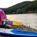 A purple camping tent set up on a sandy lakeside shore at dawn or dusk, with an inflatable stand-up paddleboard (SUP) and camping gear laid out nearby, ready for water activities and outdoor adventure.