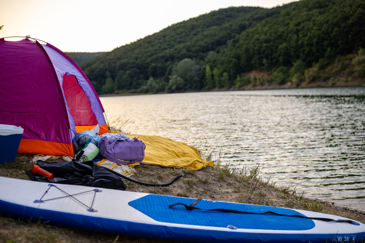 A purple camping tent set up on a sandy lakeside shore at dawn or dusk, with an inflatable stand-up paddleboard (SUP) and camping gear laid out nearby, ready for water activities and outdoor adventure.