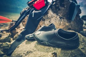 A pair of black water shoes sits on a rocky shore, with a wetsuit, a red dry bag, and an orange paddle partially visible in the background. This gear is ready for water sports and provides protection and comfort in wet outdoor environments.