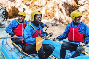 hree individuals wearing yellow helmets, blue shirts, and red life vests sit in an inflatable blue raft, holding paddles. They appear to be whitewater rafting or enjoying a similar water adventure on a river, with rocky terrain in the background.