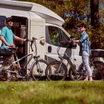 Two people stand with their bicycles beside a white camper van, parked on green grass at a sunny campground. This image represents convenient personal transport options, like e-bikes, for exploring while camping.