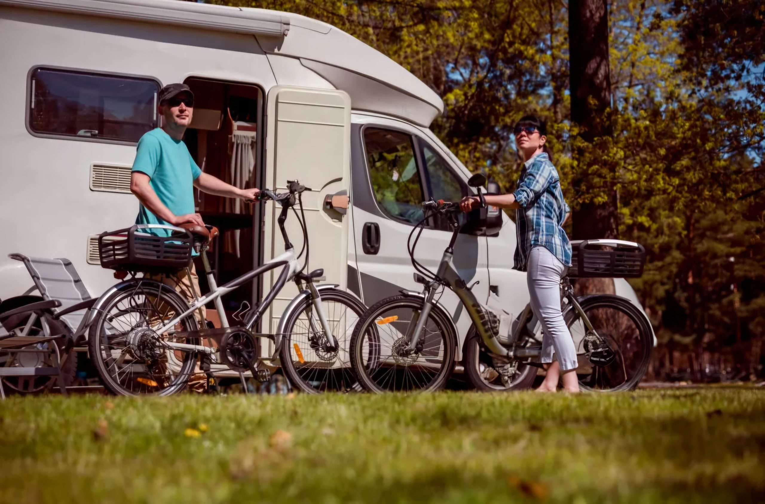 Two people stand with their bicycles beside a white camper van, parked on green grass at a sunny campground. This image represents convenient personal transport options, like e-bikes, for exploring while camping.