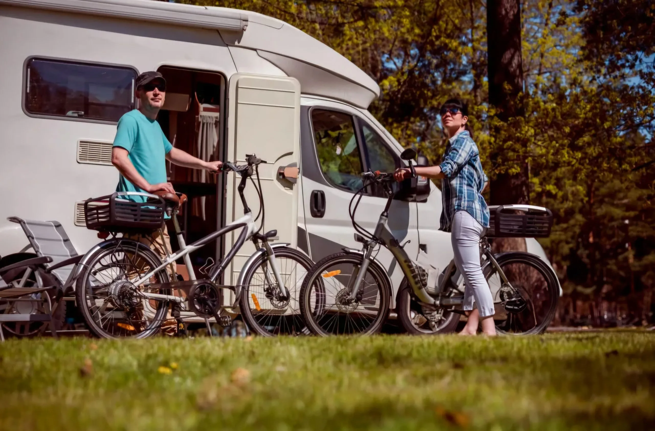 Two people stand with their bicycles beside a white camper van, parked on green grass at a sunny campground. This image represents convenient personal transport options, like e-bikes, for exploring while camping.