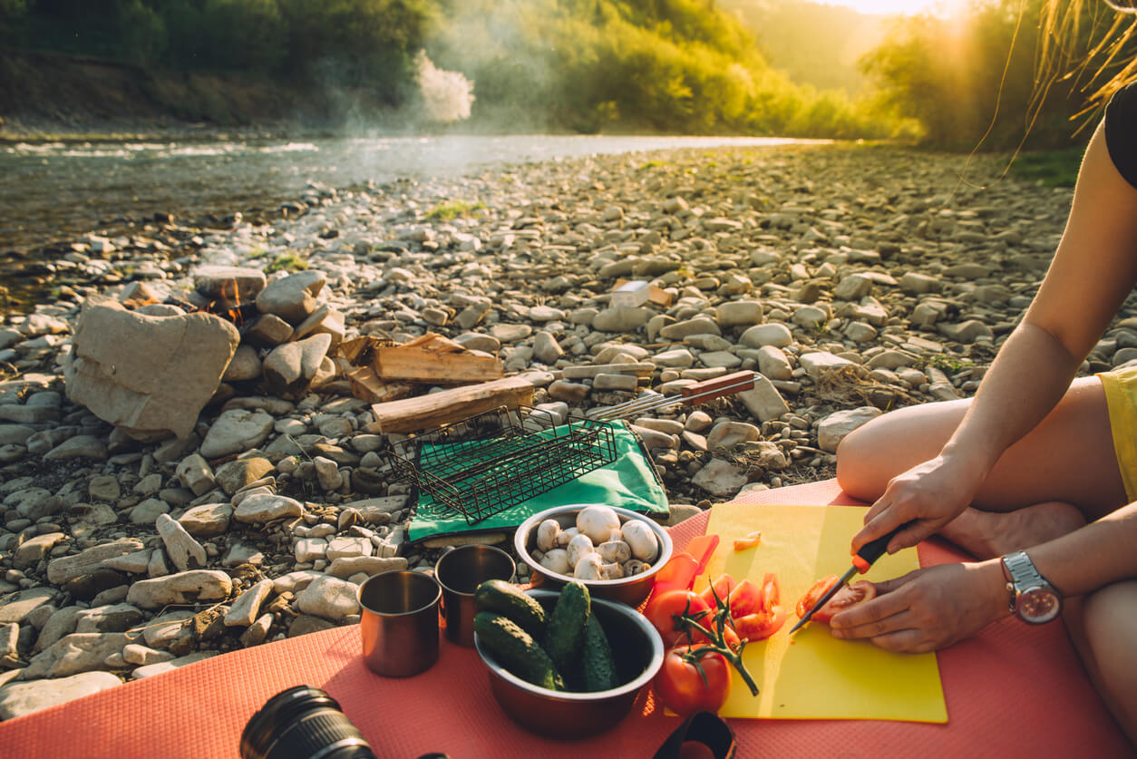 A person sits on a mat on a rocky riverbank, preparing fresh vegetables like tomatoes, cucumbers, and mushrooms on a cutting board, with metal camping cups and a grill grate nearby. In the background, a small campfire smokes beside a flowing river with sunlit green hills, depicting easy outdoor meal preparation while camping.
