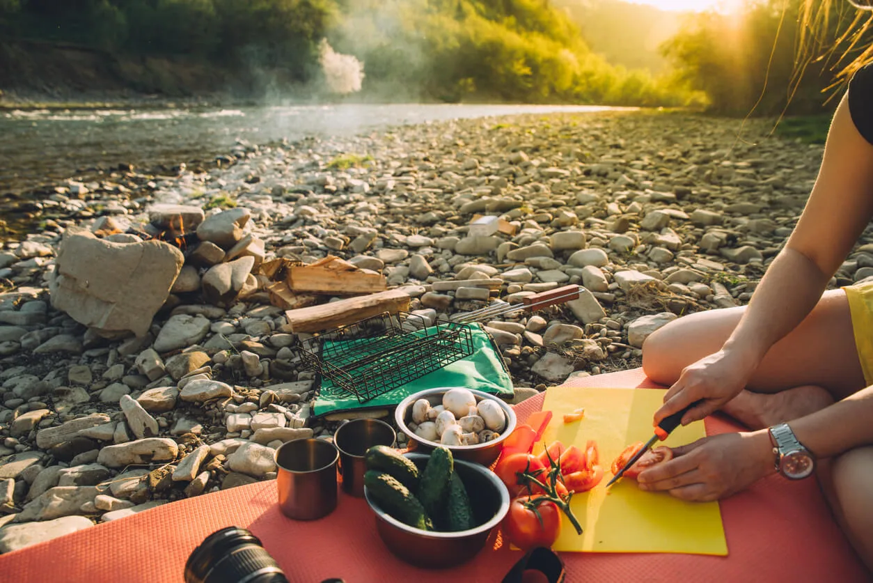 A person sits on a mat on a rocky riverbank, preparing fresh vegetables like tomatoes, cucumbers, and mushrooms on a cutting board, with metal camping cups and a grill grate nearby. In the background, a small campfire smokes beside a flowing river with sunlit green hills, depicting easy outdoor meal preparation while camping.