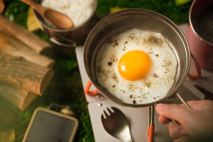 A perfectly fried egg with a bright yellow yolk and sprinkled pepper cooks in a small metal pan on a portable camping stove, with a bowl of rice and other camping gear visible in the background, illustrating an easy outdoor breakfast.