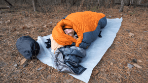 Man in an orange sleeping bag with a sheet underneath it in the middle of a clearing waking up.