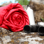Red rolled up sleeping bag next to some binoculars and a water tin bottle on a rock.