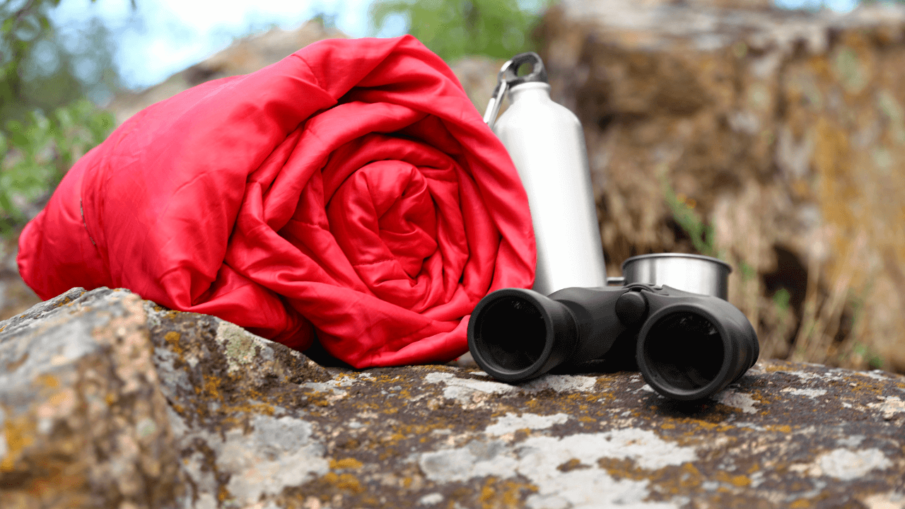 Red rolled up sleeping bag next to some binoculars and a water tin bottle on a rock.