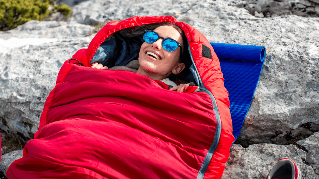 Woman with shades completely snug in a red sleeping bag, smiling.