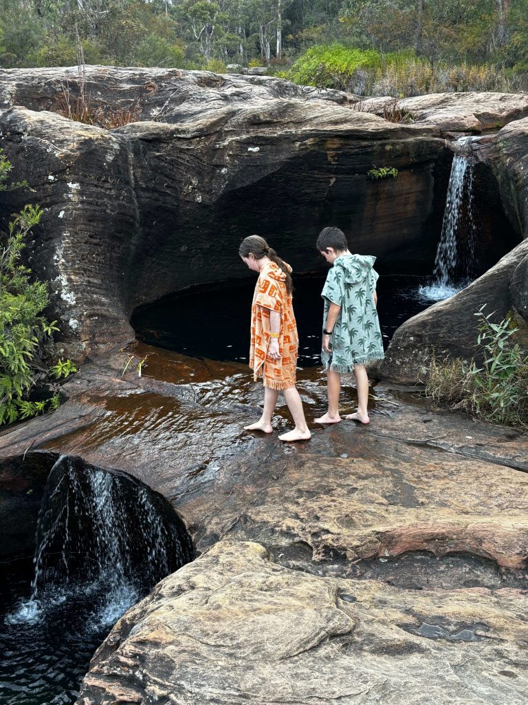 Two barefoot "wildlings," Australian children, traverse dark, wet rocks near a small waterfall. The girl on the left wears an orange hooded towel, and the boy on the right wears a green patterned hooded beach towel. These fun hooded towels for kids are perfect for a kids' beach holiday or family beach day in Australia.